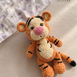 May include: A plush crochet tiger toy with orange and cream yarn, a pink nose, and black embroidered stripes. The toy features round ears, a rounded snout, and a long tail with a black tip. The text PDF crochet pattern is visible.