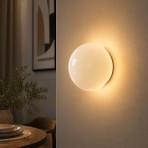 Könnte beinhalten: Eine kreisförmige Wandleuchte mit einer glatten, opaken weißen Glaskugel, die ein warmes, sanftes Licht ausstrahlt. Die kugelförmige Lampe ist direkt an einer flachen, neutralfarbenen Wand befestigt und erzeugt einen diffusen Lichteffekt.