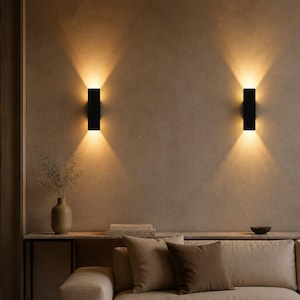 May include: Two rectangular black wall sconces mounted on a textured beige wall, each emitting a warm, dual-directional light beam that casts a soft glow upwards and downwards, creating a symmetrical illumination pattern in the room.