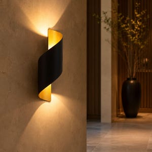 Puede incluir: Un aplique de pared con un diseño metálico curvo en forma de S, que presenta un exterior negro mate y un interior dorado reflectante. La lámpara emite una luz cálida y brillante desde las aberturas superior e inferior hacia la pared.