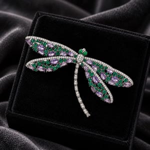 Puede incluir: Un broche de libélula con alas incrustadas de piedras ovaladas de color lavanda y piedras redondas verdes, bordeadas por cristales blancos. El cuerpo y la cola están detallados con pequeños cristales transparentes y dos piedras verdes en la cabeza.