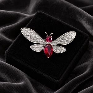 Puede incluir: Un broche con forma de insecto en tono plateado que presenta dos piedras rojas facetadas en la cabeza y el abdomen. Las alas tienen un patrón de filigrana detallado y están incrustadas con numerosas piedras pequeñas y claras.
