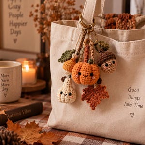 May include: A beige canvas tote bag with the text Good Things Take Time and a heart symbol, featuring a metal keyring attached to the handle with several small crocheted charms including an orange pumpkin, a white pear, an acorn, and a leaf.