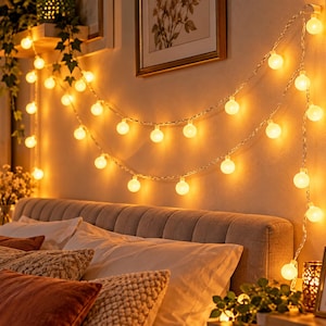 Puede incluir: Una cadena de luces LED con forma de globo que emiten un cálido resplandor amarillo, dispuestas en dos bucles horizontales sobre una pared encima de un cabecero capitoné. Las luces están conectadas por un cable blanco fino y flexible.