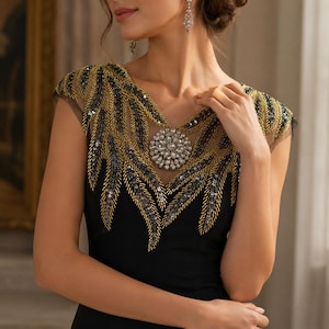 Puede incluir: Un vestido negro sin mangas con un panel transparente en el escote, decorado con bordados de lentejuelas doradas y plateadas en forma de hojas. Un gran racimo circular de cristales transparentes se sitúa en el centro del escote.