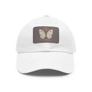 Op de afbeelding: Een witte baseballpet met een rechthoekige donkergrijze patch aan de voorzijde. De patch toont een gedetailleerde vlinderillustratie met ingewikkelde bloemmotieven in gedempte tinten en de tekst Fly High in een sierlijk lettertype.