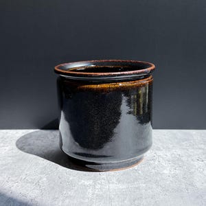 May include: A cylindrical ceramic planter with a glossy black glaze and a dark brown rim. The vessel features a smooth, reflective surface and a slightly tapered base, showcasing a deep, dark finish that contrasts with the warm brown edge.