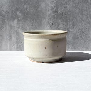 May include: A small, off-white ceramic bowl with a rounded body and a slightly flared rim. The bowl has a simple, minimalist design and sits on a white surface. The background is a textured gray wall. The bowl is approximately 3 inches in diameter.