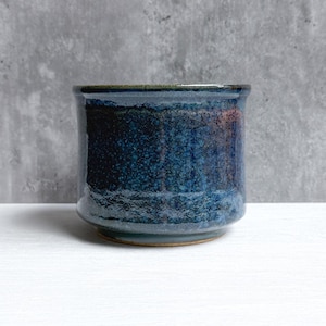 May include: A cylindrical ceramic cup featuring a deep blue glaze with mottled textures and subtle hints of brown and black. The vessel has a smooth, slightly flared rim and a solid base, showcasing a reactive finish that creates a unique depth of color across the exterior surface of the piece.