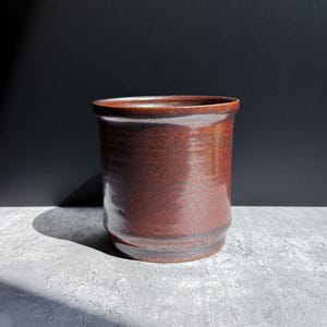 May include: A cylindrical ceramic vessel with a deep reddish-brown glaze and a subtle speckled texture. The pot features a slightly flared rim and a stepped base design, showcasing a smooth, reflective surface finish throughout the exterior.