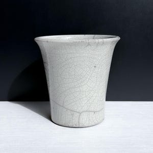 May include: A ceramic cup featuring a light gray glaze with a prominent dark crackle pattern throughout the surface. The vessel has a flared rim, a cylindrical body that tapers slightly toward the base, and a smooth, uniform finish.