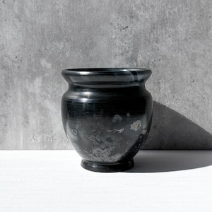 May include: A black ceramic vase with a rounded body and a slightly flared rim. The vase has a glossy finish with subtle variations in color and texture. The vase sits on a white surface against a gray background.