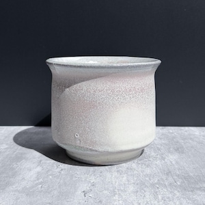 May include: A ceramic planter with a flared rim and a tapered base. The exterior features a speckled glaze with a gradient transition from a soft, muted pink at the top to a creamy off-white at the bottom, showcasing a smooth, matte finish.