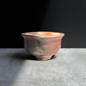 May include: A small, round ceramic bowl with a flared rim and three short feet. The exterior features a textured, mottled finish with a gradient of warm orange, reddish-brown, and cream tones, transitioning into a speckled, rough-looking surface.