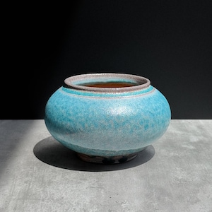 May include: A round ceramic bowl with a wide, bulbous body and a short, narrow neck. The exterior features a textured, mottled glaze in shades of bright turquoise and light blue, while the rim shows a subtle, earthy brown and grey tone.