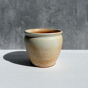 May include: A handcrafted, round ceramic pot with a wide opening. The pot has a speckled, off-white exterior with warm brown tones near the rim and base. The pot sits on a white surface against a gray backdrop.