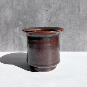 May include: A dark brown ceramic vase with a wide rim and a cylindrical body. The vase has a speckled texture and sits on a white surface. The background is a gray wall.