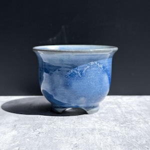 May include: A small, round ceramic planter featuring a variegated blue and white glaze with a smooth, glossy finish. The vessel has a flared rim and is supported by three short, integrated feet, showcasing a blend of deep and light blue tones.