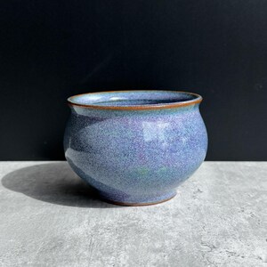 May include: A small, rounded ceramic bowl with a speckled blue and purple glaze. The rim and base feature a thin, exposed brown clay edge, contrasting with the smooth, glossy finish of the main body of the vessel.