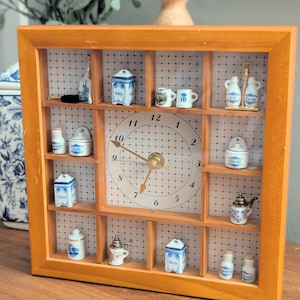 May include: A square wooden wall clock with a grid-patterned face featuring numbers 1 through 12. The clock frame contains multiple small compartments displaying miniature ceramic kitchen canisters, jars, and jugs with blue and white patterns.