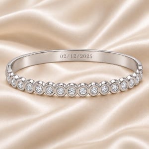 May include: A silver-toned hinged bangle bracelet featuring a row of round, clear stones set in individual circular bezels. The inner surface of the metal band is engraved with the date 02/12/2025 in a clean, sans-serif font style.