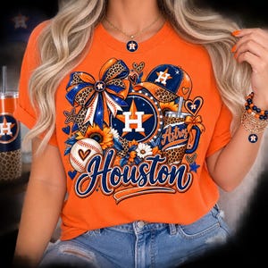 May include: An orange short-sleeved t-shirt featuring a graphic print with a Houston Astros logo, a baseball, a leopard print bow, a baseball cap, sunflowers, and the word Houston in blue script. The design includes hearts and stars.