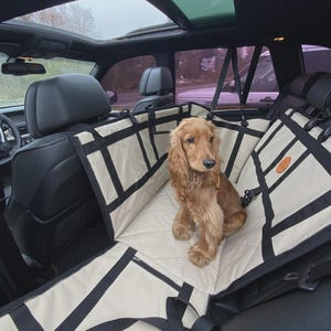May include: A beige quilted car seat cover for pets with black nylon straps and buckles, installed across the back seat of a vehicle. A golden-brown spaniel sits on the protective mat, which features a diamond-stitched pattern and a small circular logo.