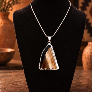 May include: A necklace featuring a triangular pendant with a polished, banded agate stone in shades of black, brown, and cream. The stone is encased in a textured silver-toned metal setting and hangs from a thin, smooth silver-toned snake chain.