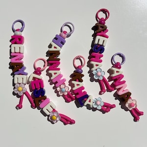 May include: Five colourful beaded keychains featuring pink nylon cords and plastic lobster clasps. Each keychain displays a name spelled out in block letters, including RENAYE, LESLIE, HANNAH, KAHLA, AYESHA, and CANNA, accented with small flower beads.