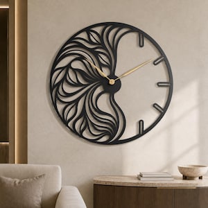 May include: A circular wall clock featuring a black metal frame with an intricate, openwork leaf-like pattern on the left side. The right side is open, with simple black hour markers. The clock has two slender, gold-colored metal hands at the center.