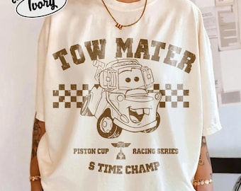 Puede incluir: Una camiseta color marfil con un gráfico marrón de una grúa de dibujos animados con el texto TOW MATER, PISTON CUP RACING SERIES y S TIME CHAMP. El diseño incluye un patrón de cuadros detrás del camión y un pequeño logotipo.