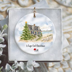 Peut inclure: Une décoration ronde en céramique blanche ornée d'une illustration à l'aquarelle d'une maison côtière avec un sapin de Noël décoré, un chemin de sable et un phare au loin. Le texte indique A Cape Cod Christmas avec l'année 2026 en dessous.