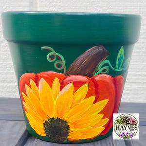 May include: A dark green plant pot featuring a hand-painted design of a bright orange pumpkin with a brown stem and green vines. A large yellow sunflower with a dark brown center is painted in front of the pumpkin. A logo reads HAYNES FAMILY FARMS.