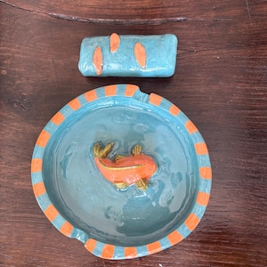 Pode incluir: Uma bandeja redonda de cerâmica com interior azul-claro apresentando um peixe koi laranja esculpido no centro e uma borda com listras alternadas em laranja e azul-claro, acompanhada por um pequeno bloco retangular azul-claro com detalhes laranja.