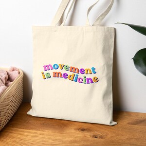 May include: A cream-colored canvas tote bag featuring the text "movement is medicine" printed in a wavy, multi-colored font. The letters are colored in shades of pink, yellow, blue, orange, and purple, with a bold, rounded, retro-inspired style.