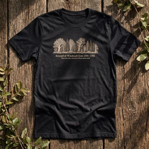Könnte beinhalten: Ein schwarzes Kurzarm-T-Shirt mit einer beigen Grafik einer Waldlinie. Unter den Bäumen steht der Text: Accused of Witchcraft from 1324 - 1782. These are only a fraction of those persecuted. Das Design ist mittig auf der Brust platziert.