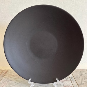 May include: A circular dinner plate with a solid matte black finish. The plate features a smooth, wide surface with a slightly concave center and a clean, unadorned rim, presenting a uniform dark appearance throughout the entire object.
