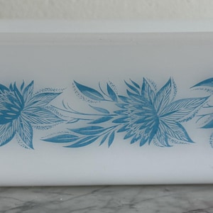 May include: A rectangular white milk glass loaf pan featuring a horizontal blue floral pattern along the side. The design consists of two stylized blue flowers with pointed petals and elongated leaves, centered on the smooth exterior of the dish.