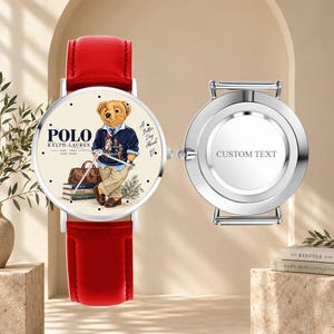 Może przedstawiać: Zegarek na rękę z jasnoczerwonym skórzanym paskiem i kremową tarczą z ilustracją misia Polo Ralph Lauren, napisem A Better Day Ahead oraz srebrnym deklem z wygrawerowanym napisem CUSTOM TEXT.
