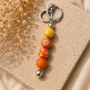 May include: A keychain featuring a silver metal ring and clasp attached to a vertical string of beads. The beads include a yellow bead with white polka dots, an orange bead with the white text hello fall, an orange bead with white polka dots, a solid orange bead, and a small silver bead at the bottom.
