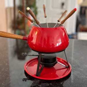 May include: A red fondue pot with a wooden handle and a set of fondue forks. The pot sits on a black metal stand with a red base. The fondue forks have wooden handles and colored tips.