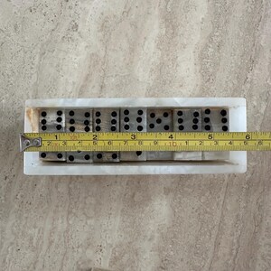 May include: A set of dominoes in a white marble tray. The dominoes are gray with black dots. A measuring tape shows the tray is approximately 6 inches long. The dominoes are arranged in a row.