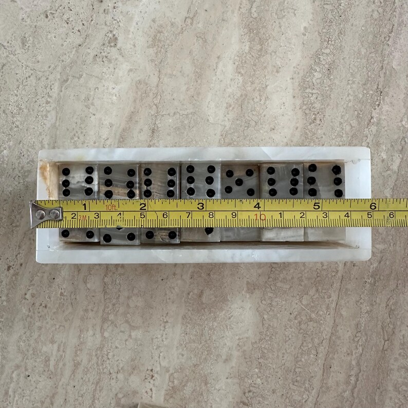 May include: A set of dominoes in a white marble tray. The dominoes are gray with black dots. A measuring tape shows the tray is approximately 6 inches long. The dominoes are arranged in a row.