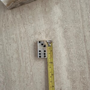 May include: A rectangular, light-colored domino with black dots, next to a measuring tape. The domino measures approximately 1 inch in width and 2 inches in length. The dots are arranged in the standard domino pattern.