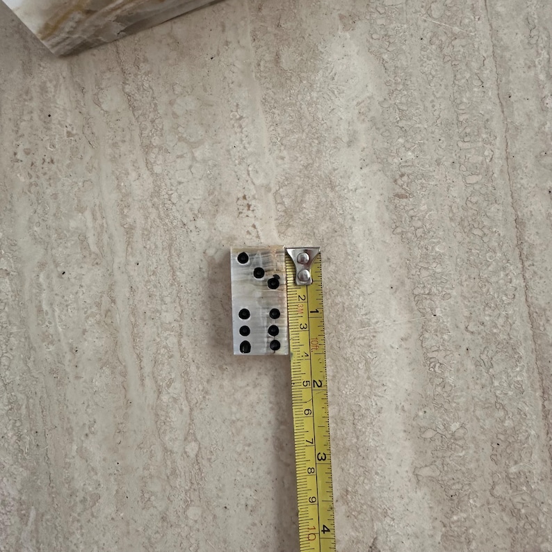 May include: A rectangular, light-colored domino with black dots, next to a measuring tape. The domino measures approximately 1 inch in width and 2 inches in length. The dots are arranged in the standard domino pattern.