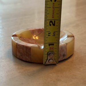 May include: A round ashtray made of marble, featuring a mix of yellow, pink, and white colors. The ashtray is approximately 1.75 inches in height. The ashtray has a classic design with a curved interior and a flat rim.