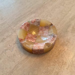 May include: A round, mosaic-style ashtray with a checkerboard pattern of beige and pink-toned stone pieces. The word "Mexico" is etched on the side. The ashtray is a decorative item.