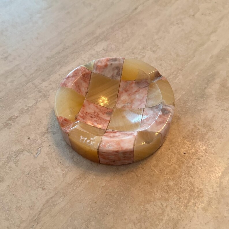 May include: A round, mosaic-style ashtray with a checkerboard pattern of beige and pink-toned stone pieces. The word "Mexico" is etched on the side. The ashtray is a decorative item.