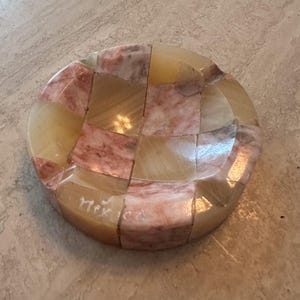 May include: A round, mosaic-style dish with a checkerboard pattern. The dish is made of polished stone in shades of beige, tan, and pink. The word "Mex" is etched on the side. The dish is likely used for decorative purposes.