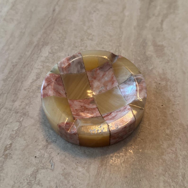 May include: A round, decorative object with a checkerboard pattern. The object is made of small, rectangular pieces in shades of yellow, pink, and brown. The surface has a glossy finish, and the object is set against a neutral background.